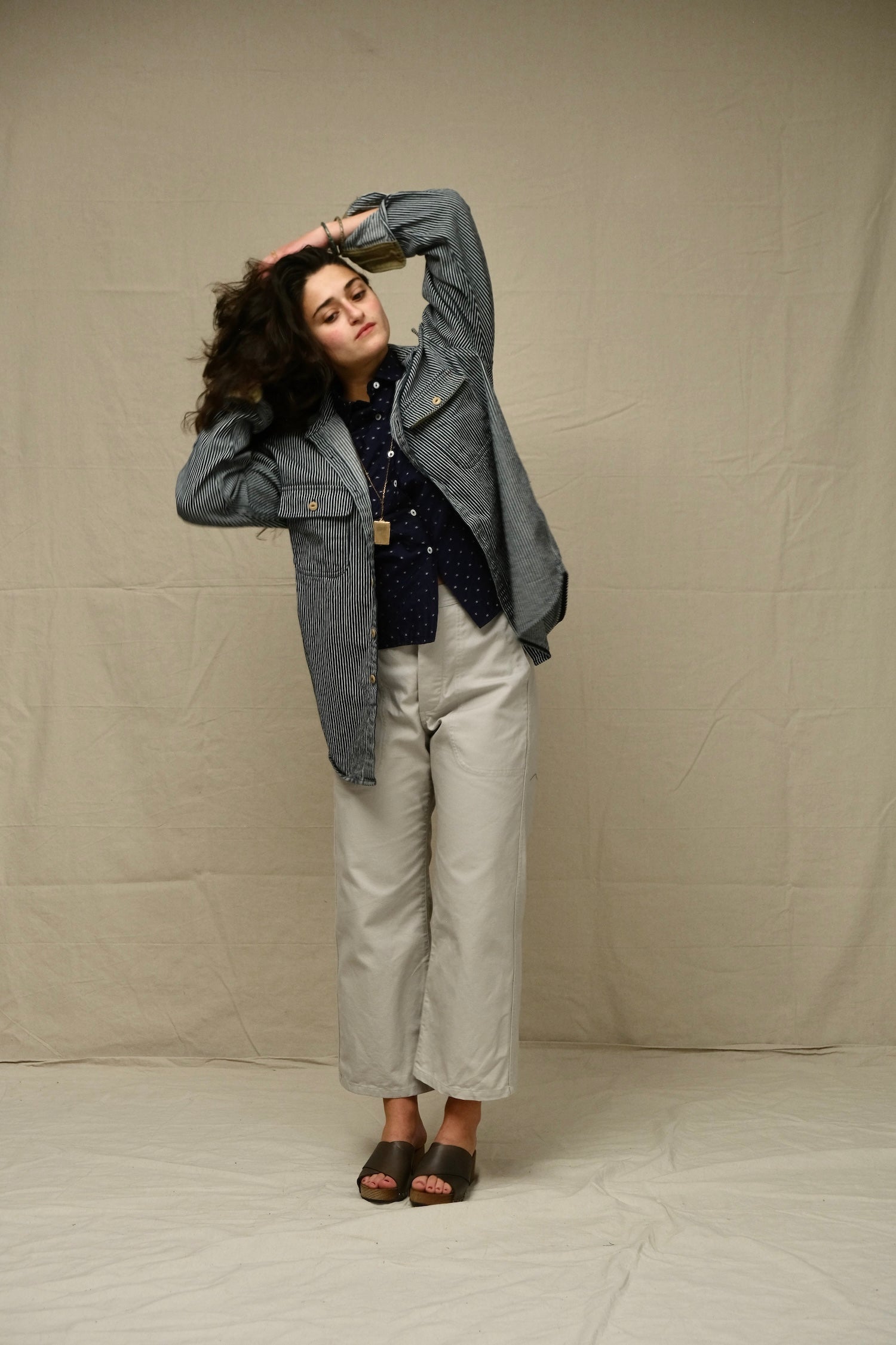 Person wearing a railroad stripe overshirt jacket and beige pants against a plain background