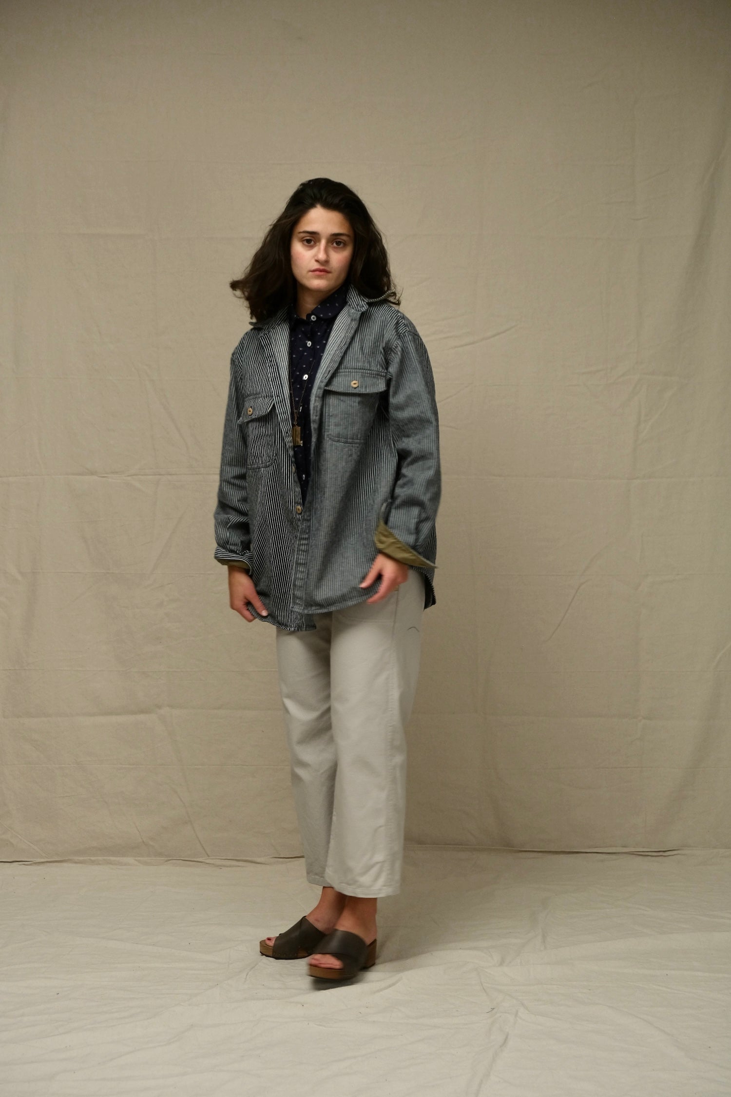 Person wearing a railroad stripe denim overshirt jacket and light-colored pants against a plain background