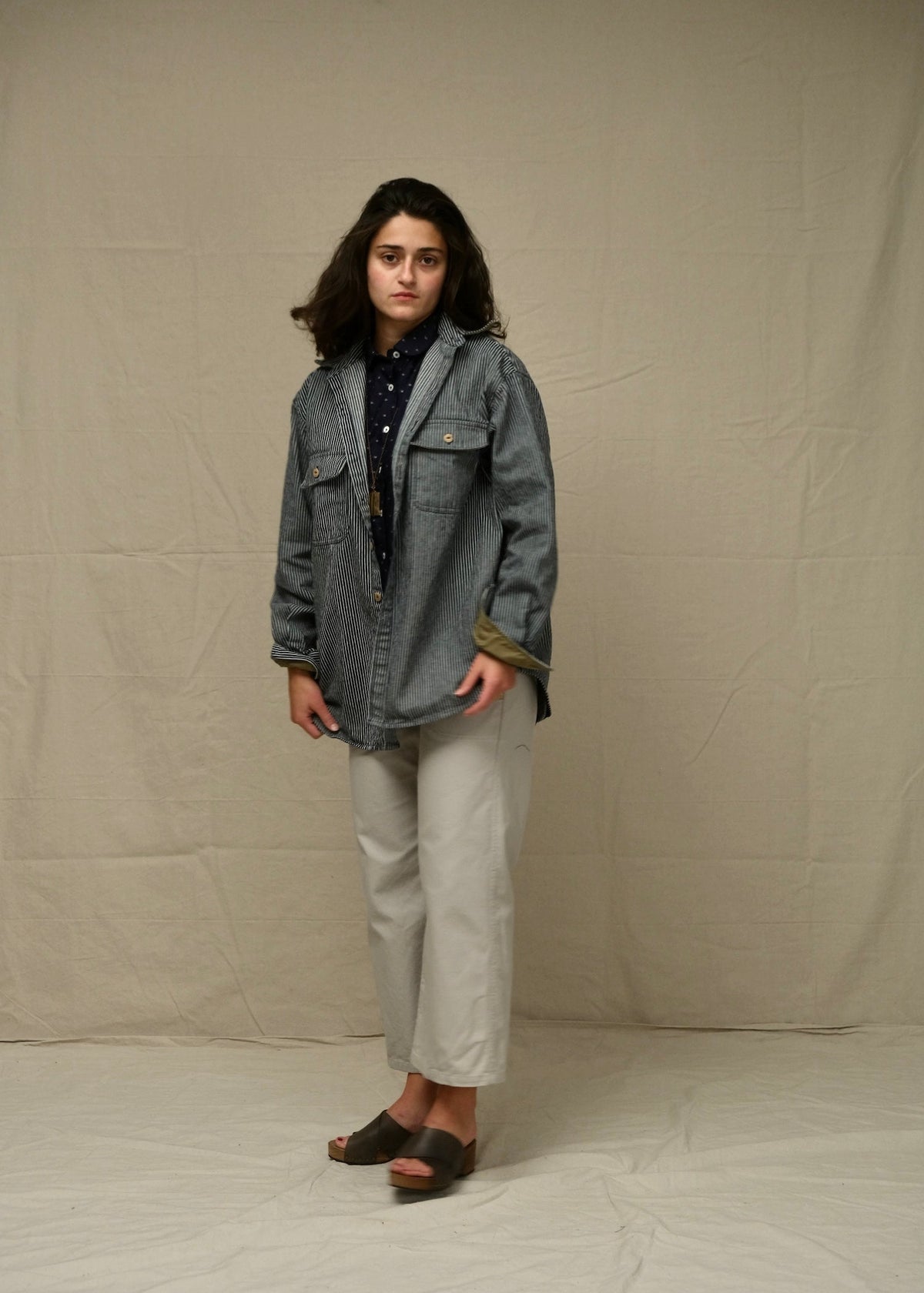 Person wearing a railroad stripe denim overshirt jacket and light-colored pants against a plain background