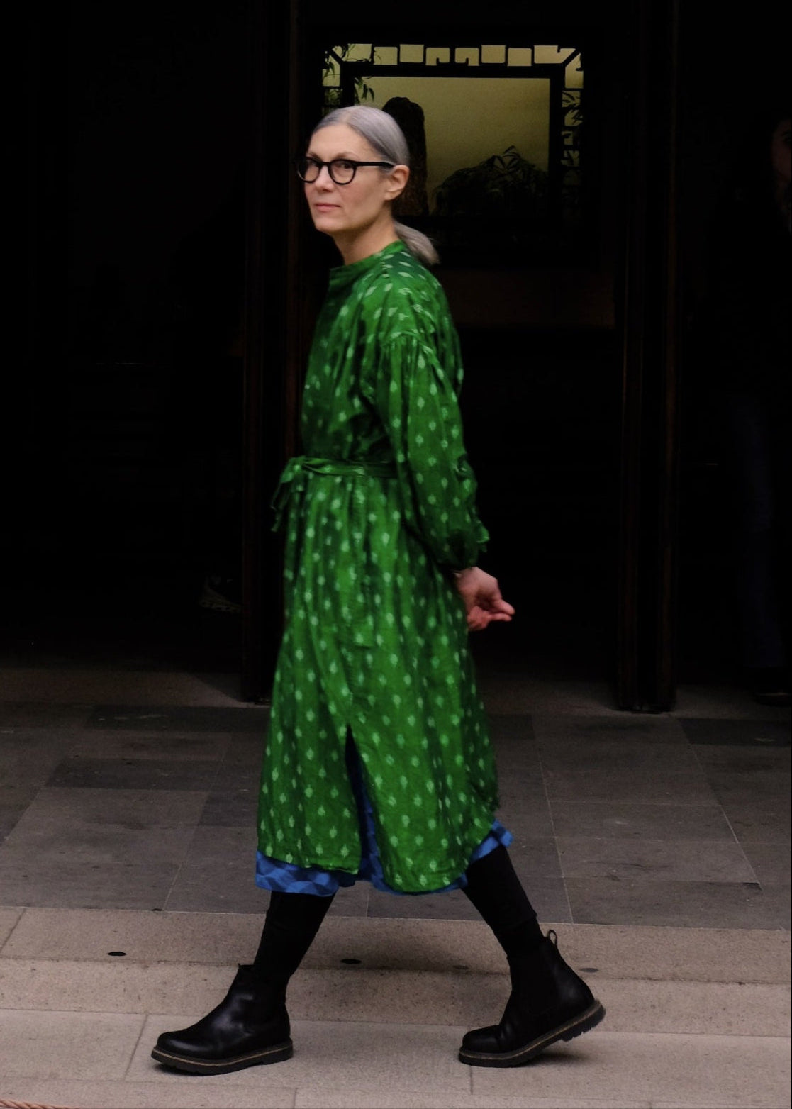 Person wearing a green patterned dress standing in front of a dark building entrance.