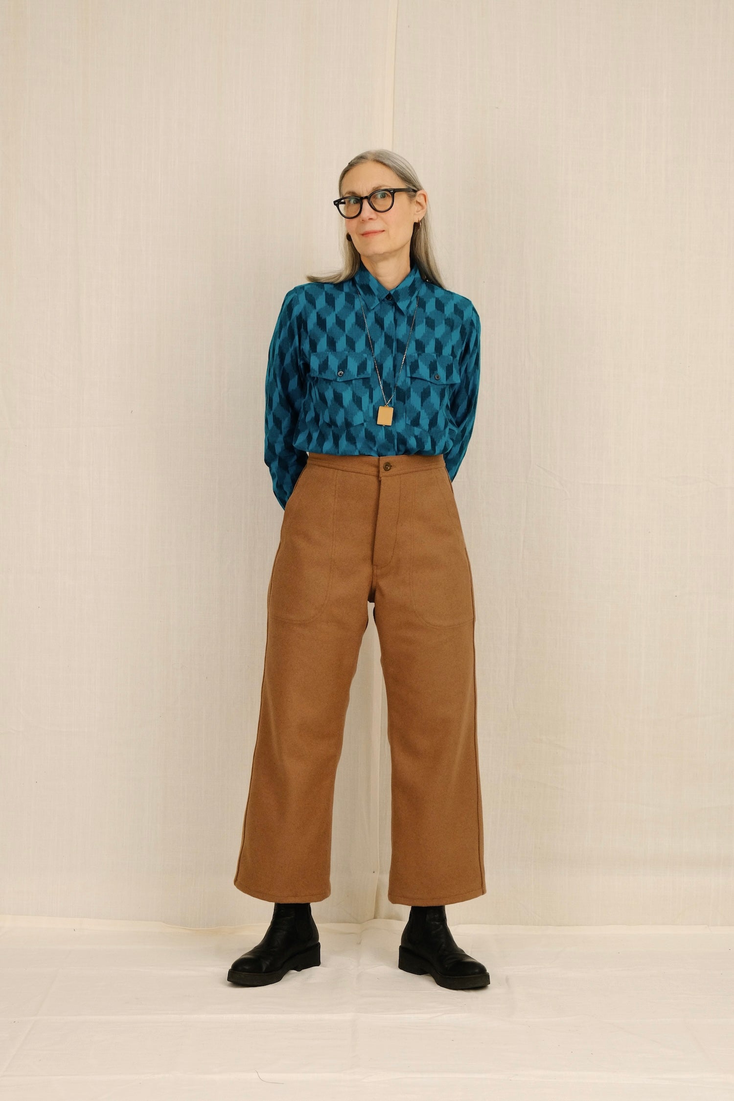 Person wearing a blue patterned shirt and brown pants against a beige background
