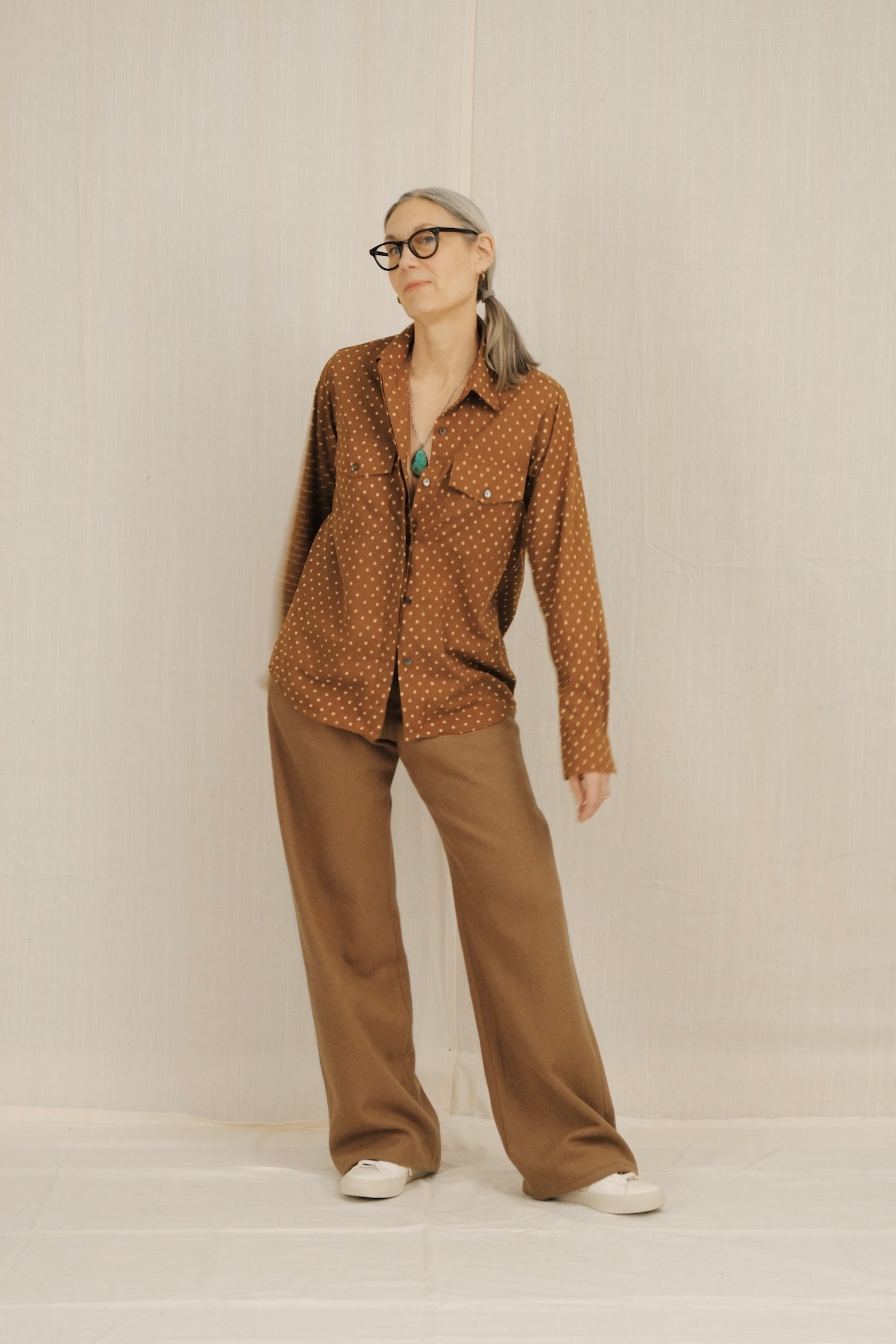 Person wearing a brown patterned shirt and beige pants against a plain background