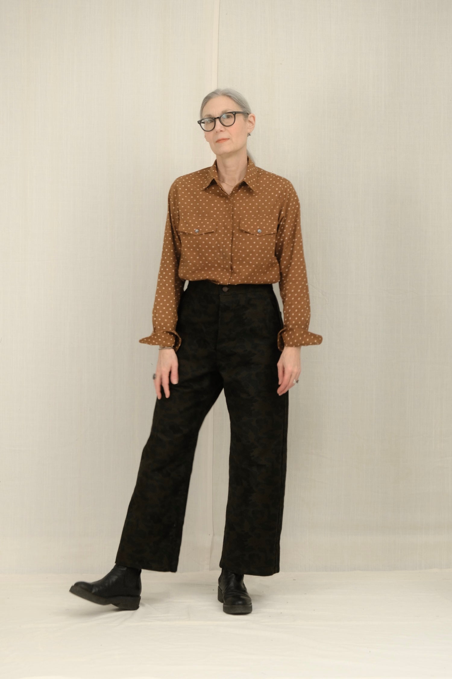 Person wearing a patterned brown shirt and black pants against a plain background