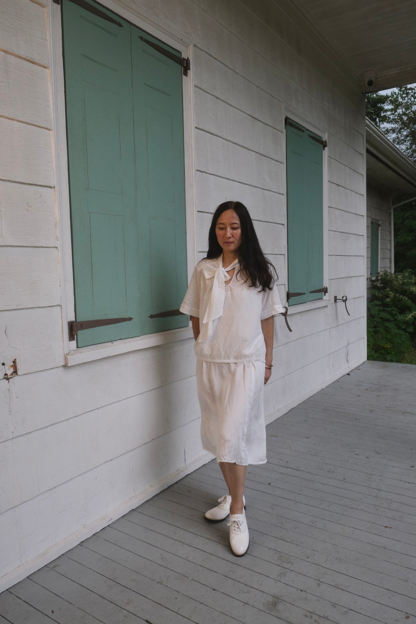 Woman in a white outfit standing on a wooden deck of a house with green shutters.