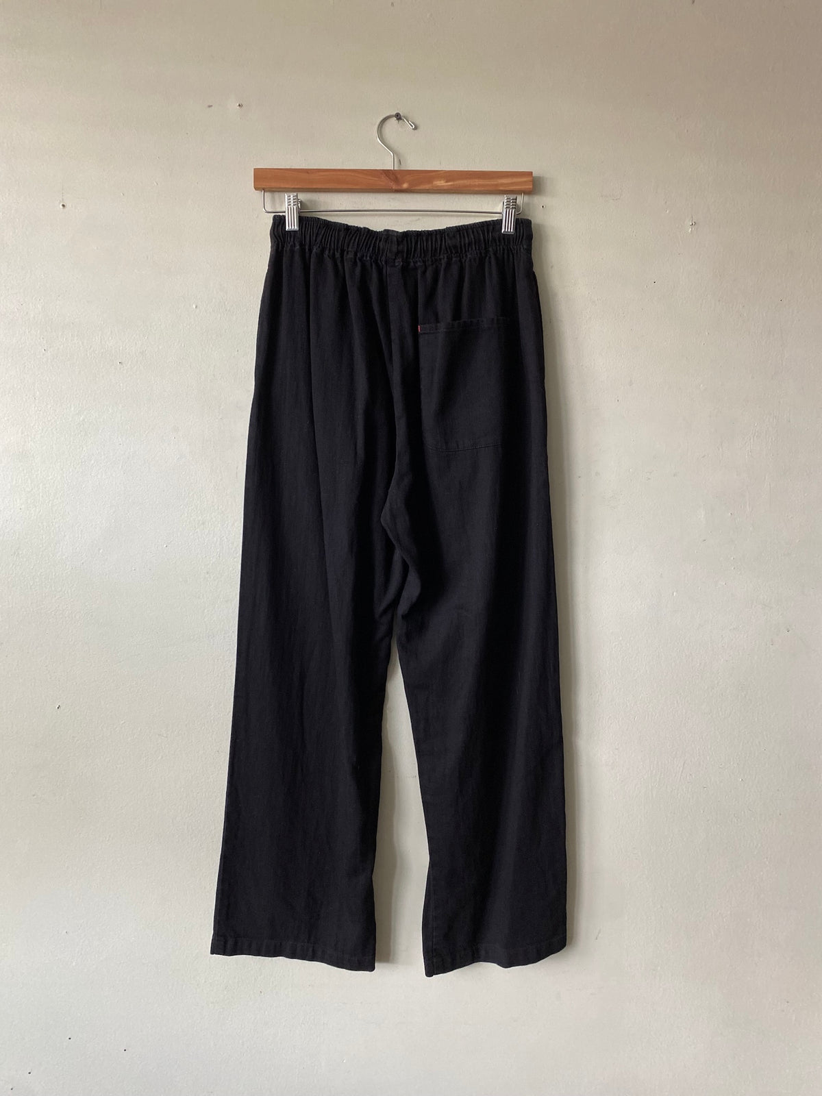 Black wide-leg cropped pants with an elastic waistband, hanging from a wooden hanger against a plain off-white wall.