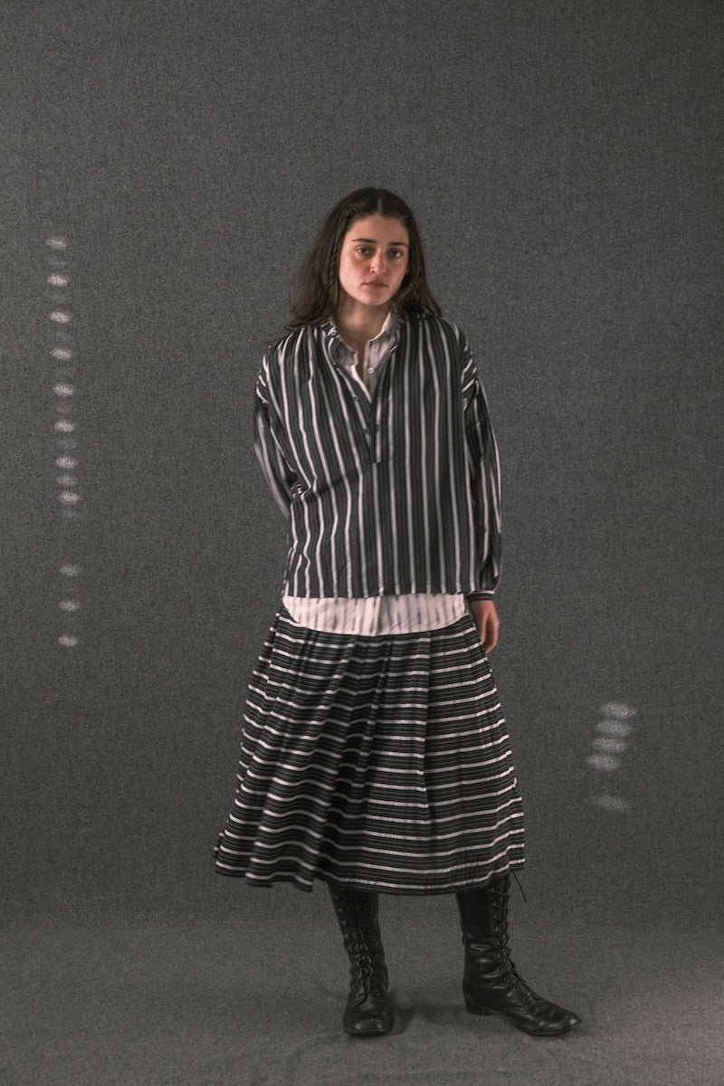 A woman stands against a dark gray studio backdrop wearing a black-and-white vertically striped long-sleeve shirt, a matching striped midi skirt, a light inner layer peeking out at the hem, and black lace-up boots.