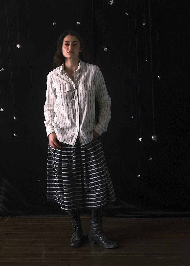 A model stands against a dark, starry backdrop wearing a white button-up shirt with thin vertical black stripes, a black and white horizontally striped midi skirt, and black leather boots.
