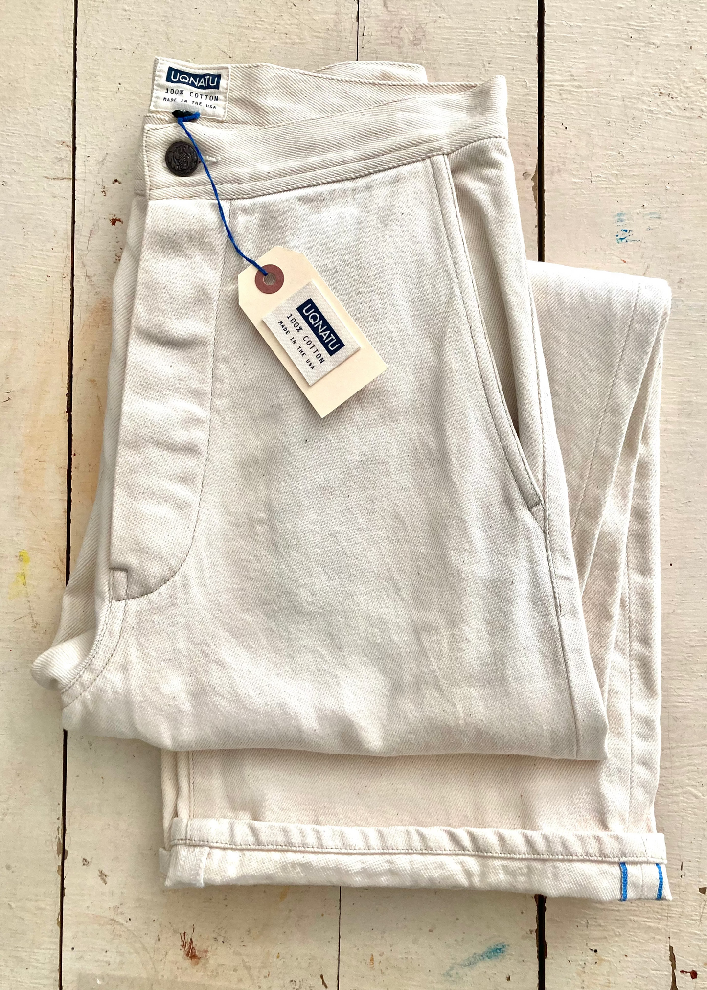 A pair of folded light beige linen pants with a visible hangtag, displayed on a rustic, light-colored wooden plank background.