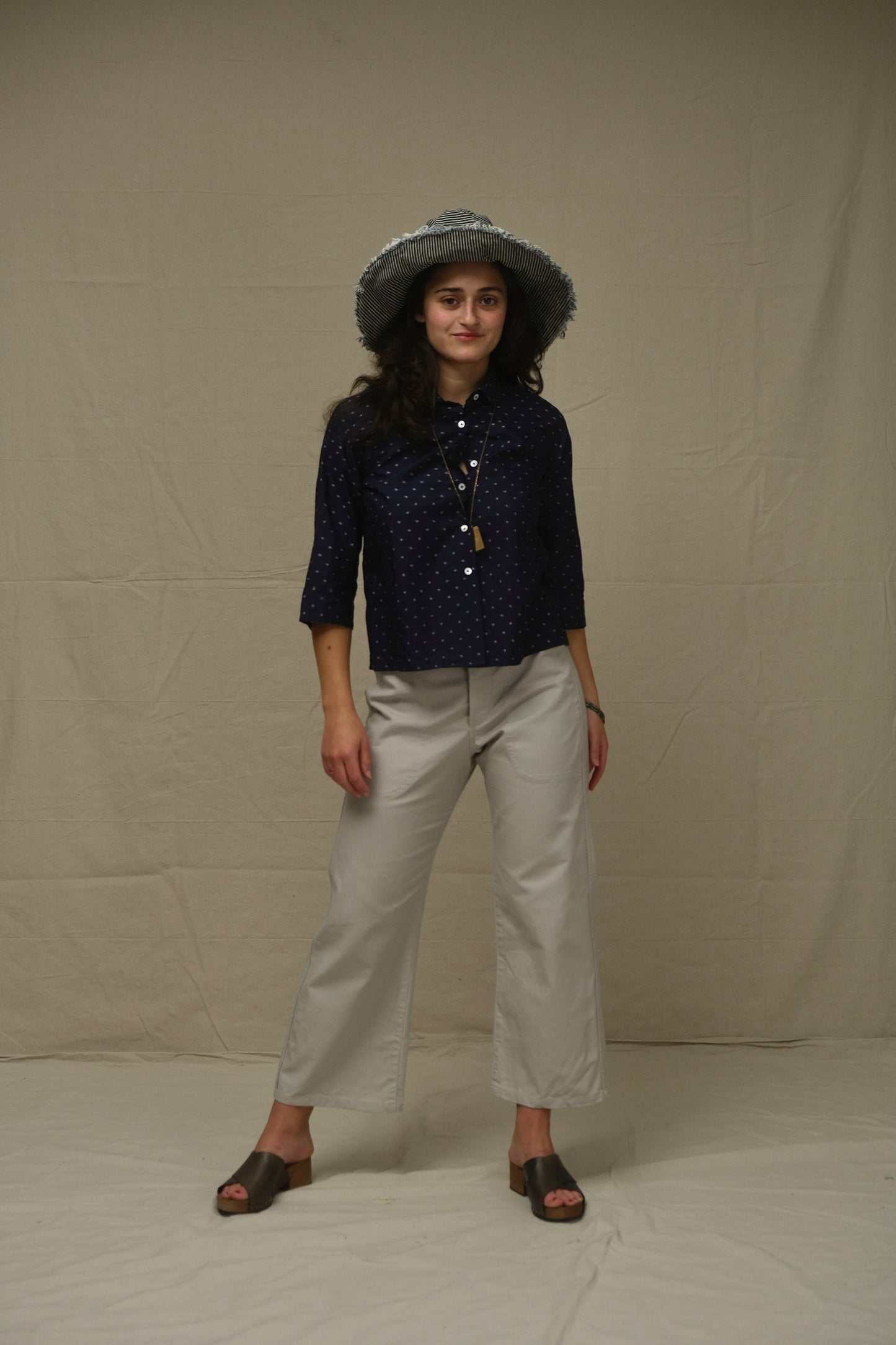 Person wearing a dark patterned shirt, light-colored pants, and a hat against a plain background