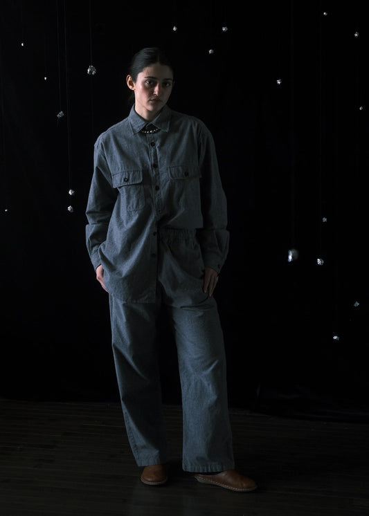 A model stands in a dark studio wearing a matching dark gray utility jumpsuit with a button-up front and wide-leg pants, paired with brown shoes against a black background with hanging light reflections.