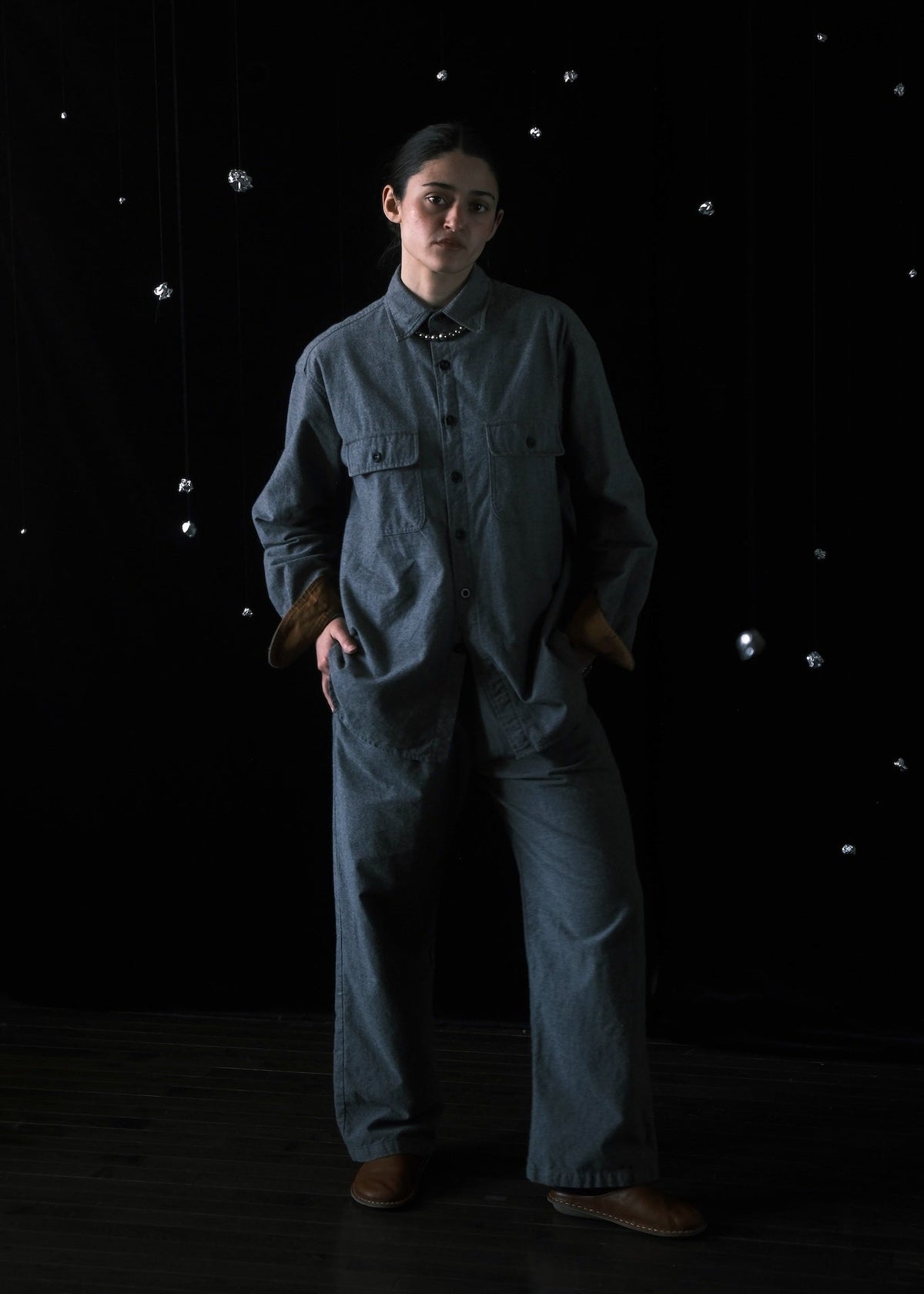 A person stands in a dark studio wearing a loose-fitting blue-gray jumpsuit or coverall with a high collar, front pockets, and wide legs, paired with brown shoes. The background is black with small scattered reflective dots.