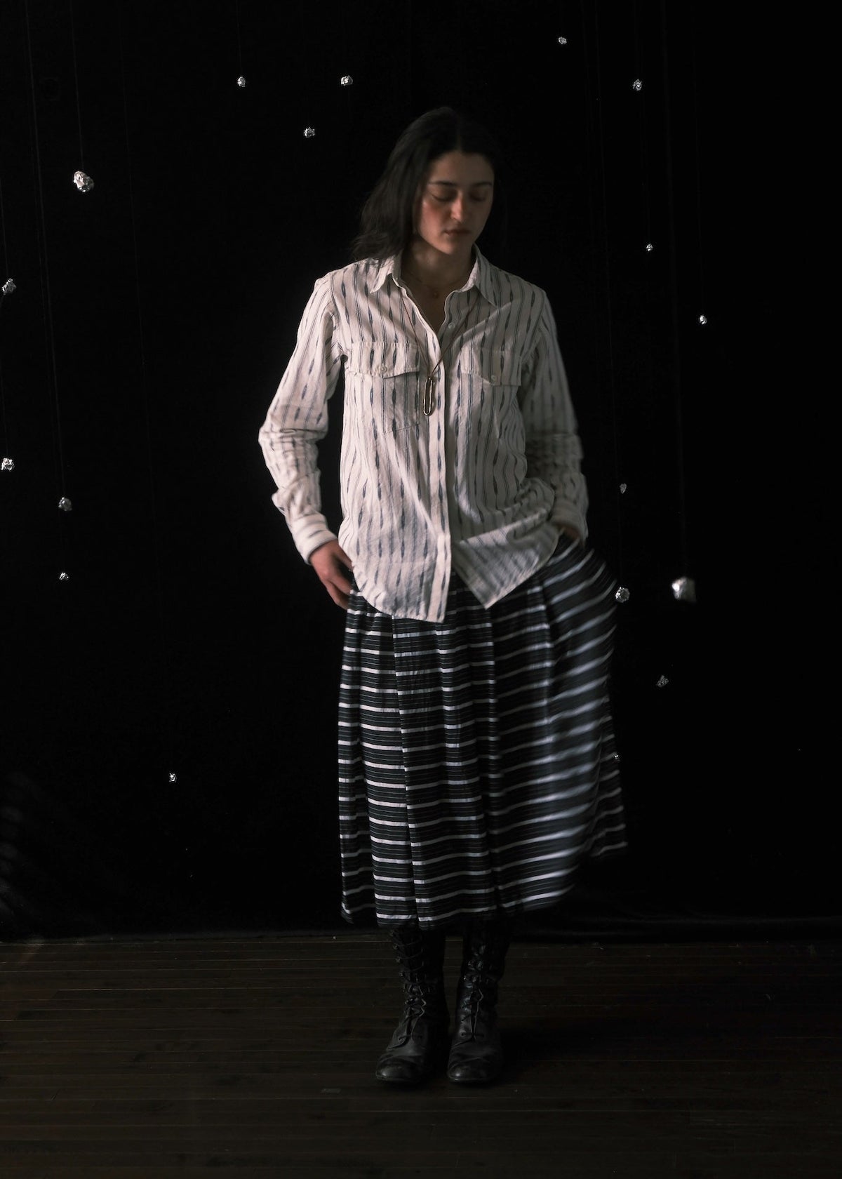A woman stands in a dark studio setting wearing a white button-up shirt with thin vertical dark stripes and a long black skirt with thin white horizontal stripes. She is wearing dark lace-up boots, and the background is black with small hanging silver reflective ornaments.