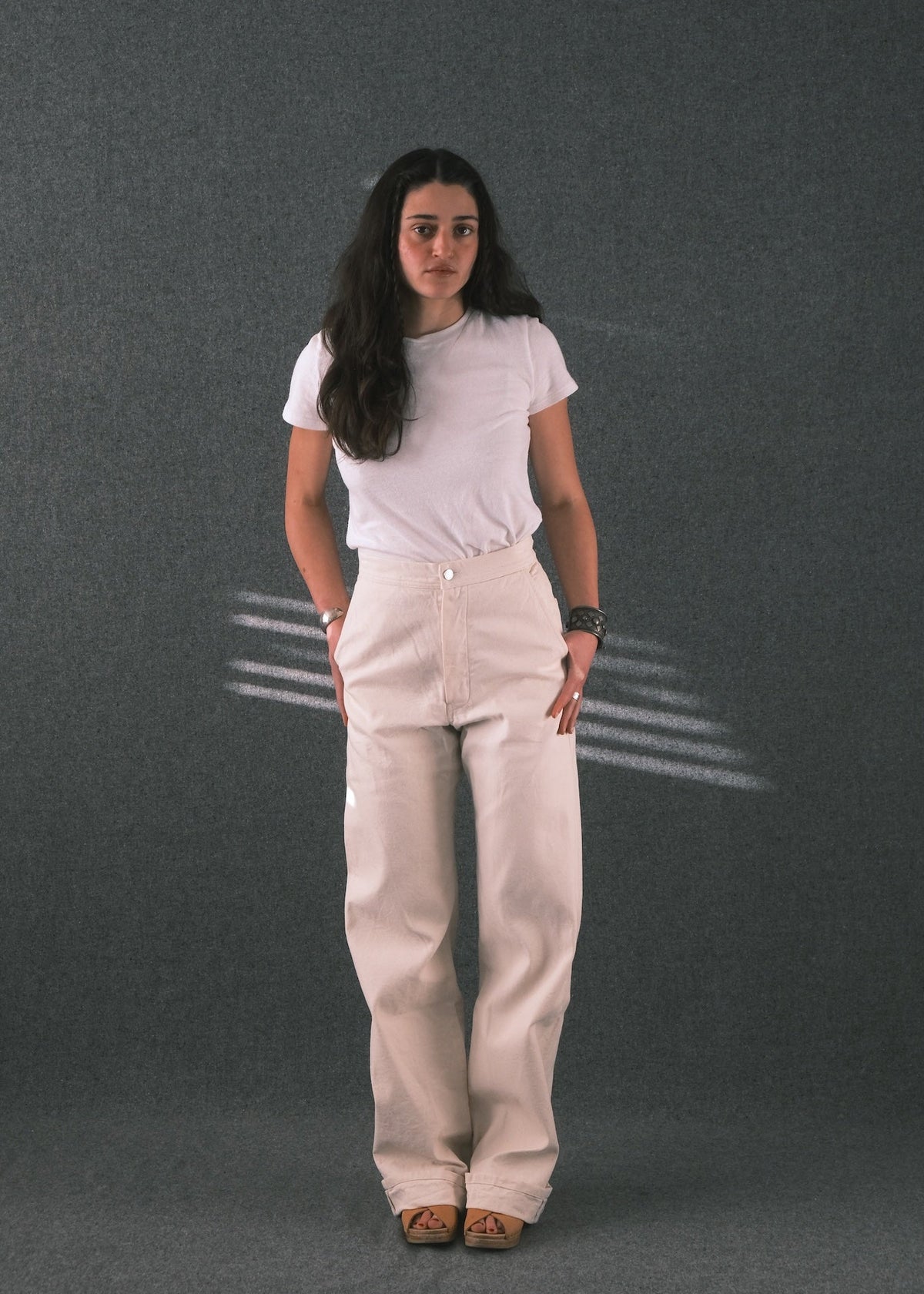 A woman stands against a dark gray textured studio backdrop wearing a white short-sleeve t-shirt and light beige wide-leg pants, paired with brown sandals.