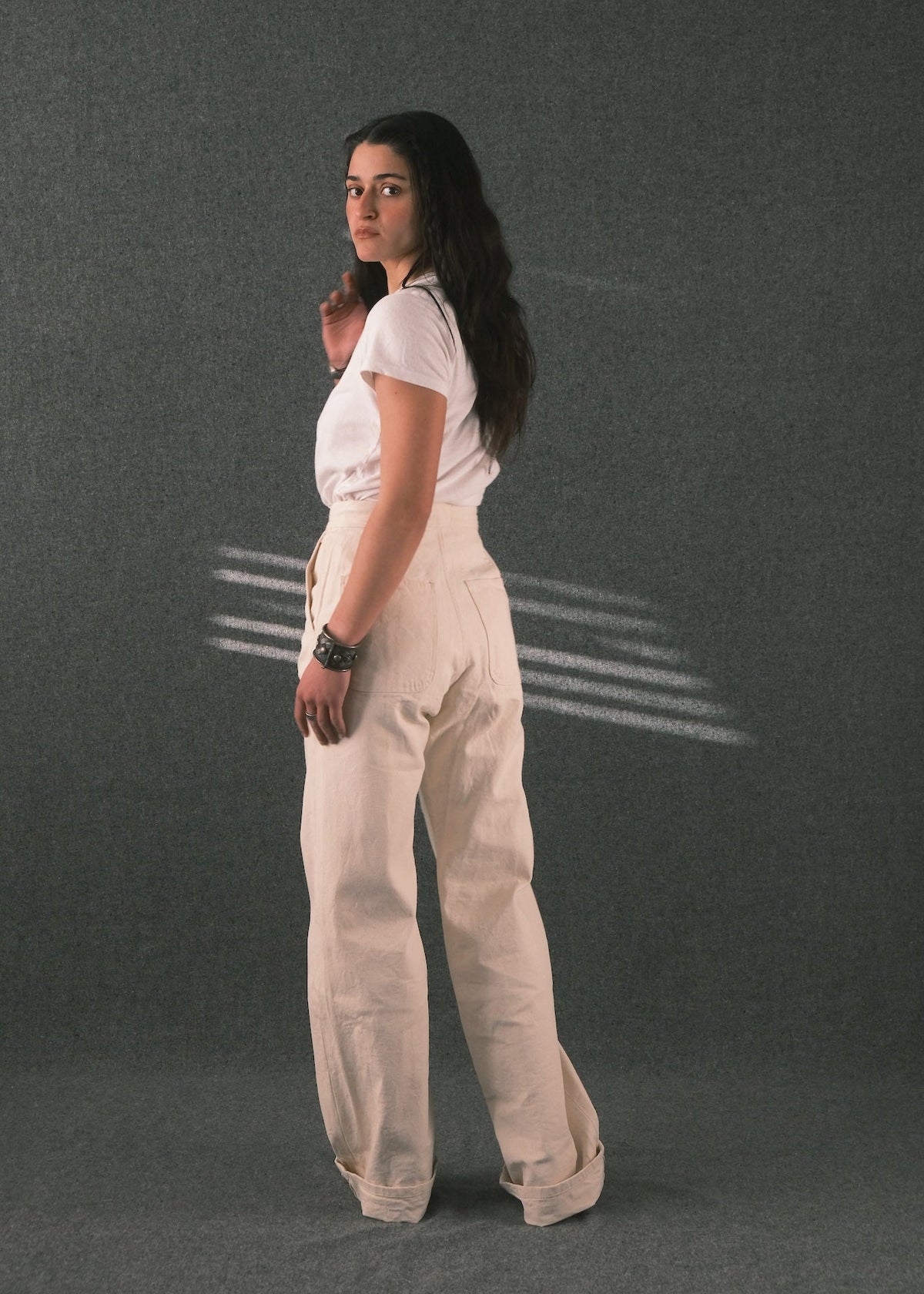 A woman stands in profile wearing a white short-sleeve top and cream wide-leg trousers against a dark gray studio backdrop with light streaks on the wall.