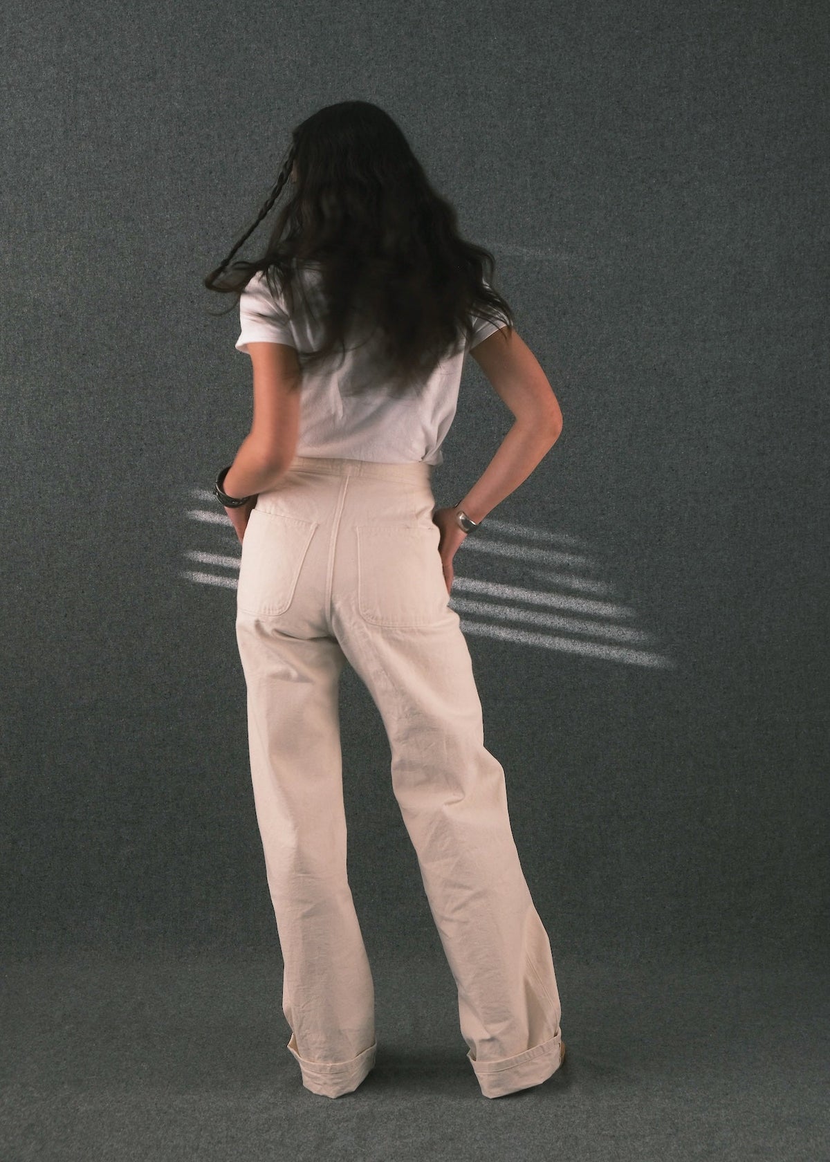 A woman is shown from behind wearing a white t-shirt and cream-colored wide-leg trousers, standing against a dark gray textured studio background.