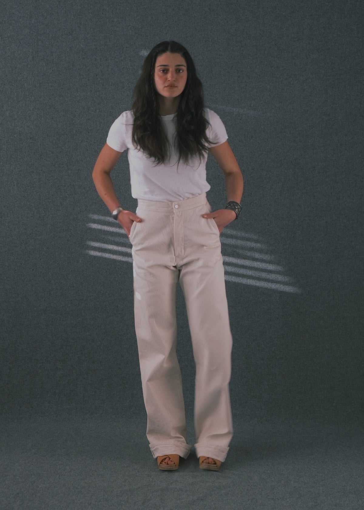 A woman stands against a textured gray backdrop wearing a white short-sleeve t-shirt and off-white wide-leg pants, with her hands in the pockets of the pants.
