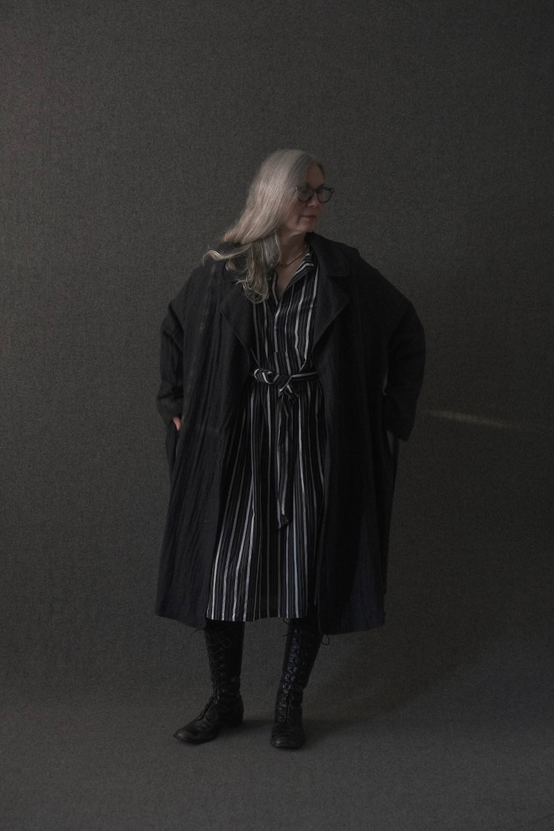 A person with long gray hair and glasses stands against a dark gray studio background wearing a long black coat over a black-and-white vertically striped shirt dress, with black lace-up boots.