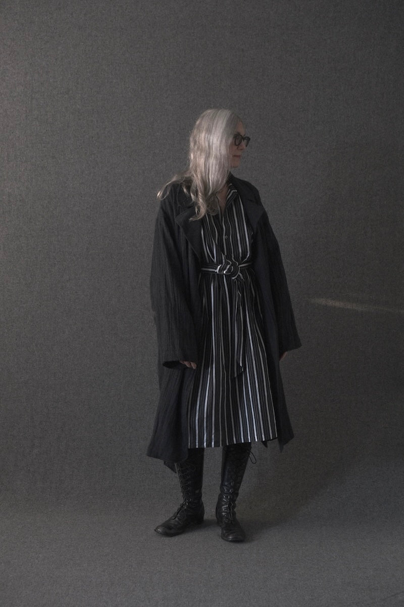 A model with long silver hair and glasses stands in profile wearing a long black belted coat over a black and white vertically striped dress, paired with black lace-up boots, against a dark gray studio backdrop.