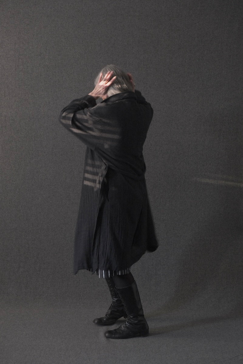 A person wearing a long black draped coat or shawl with striped detailing, standing in profile against a dark gray studio background.