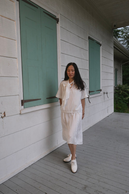Woman in a white outfit standing on a wooden deck of a house with green shutters.