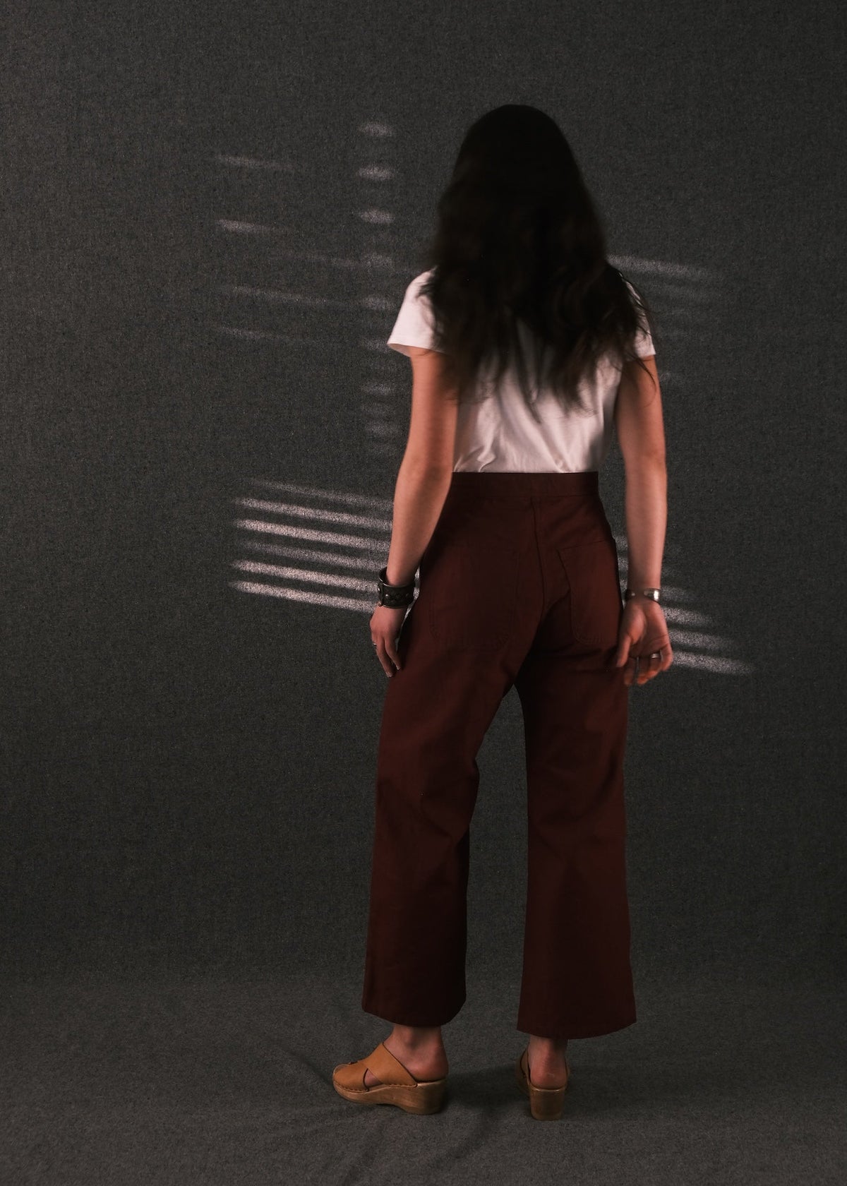 A woman is shown from behind wearing a white short-sleeve t-shirt and dark brown wide-leg cropped pants, standing against a dark gray textured backdrop with light shadows.