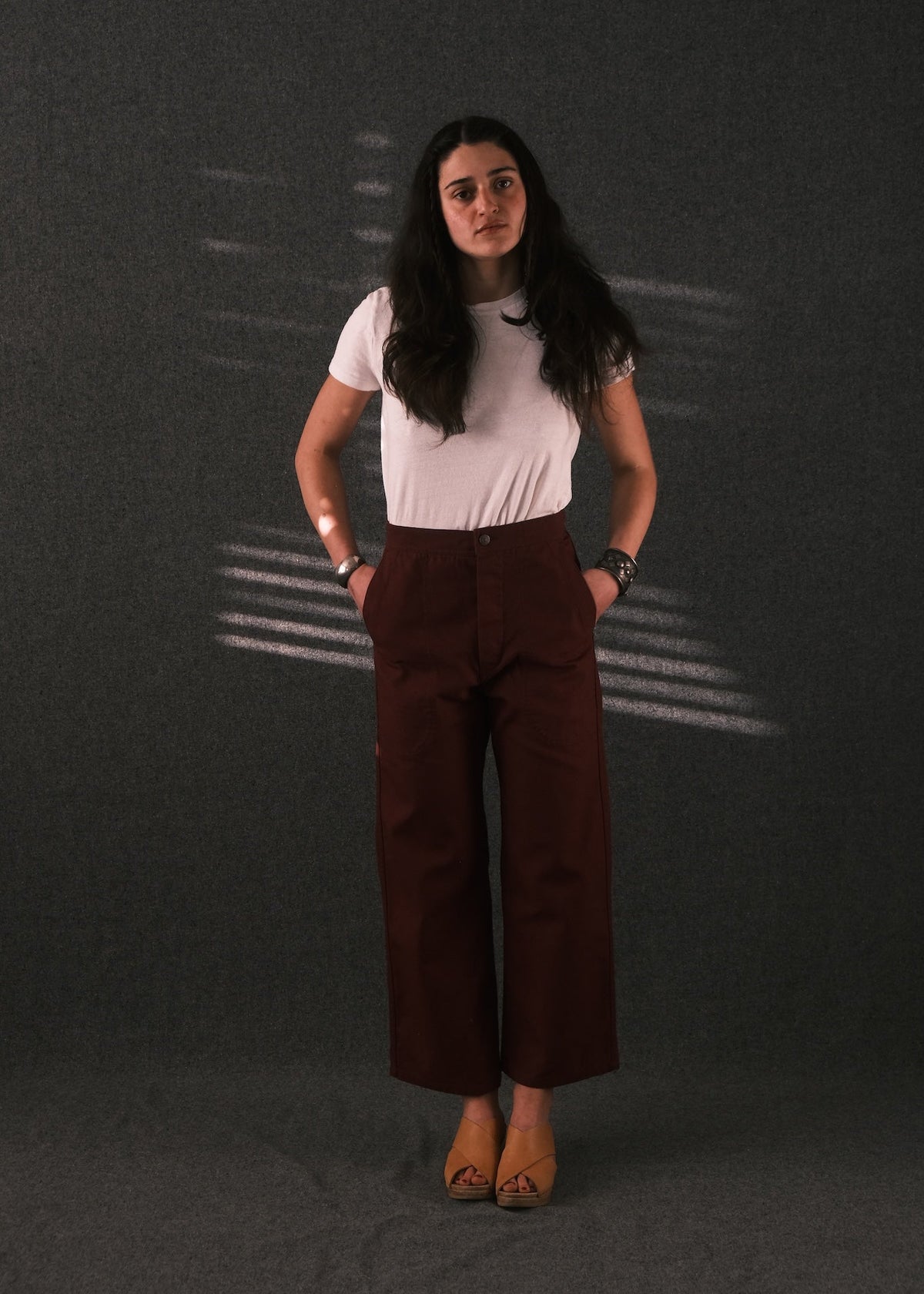 A woman stands against a dark gray textured backdrop wearing a white short-sleeve t-shirt and dark brown wide-leg cropped trousers, paired with tan flat shoes.