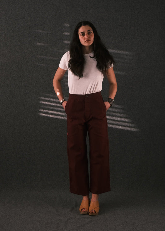 A woman stands against a dark gray textured backdrop wearing a white short-sleeve t-shirt and dark brown wide-leg cropped trousers, paired with tan flat shoes.