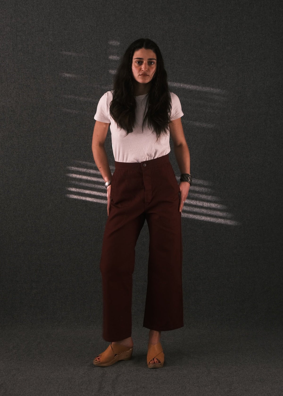 A woman stands against a dark gray textured backdrop wearing a white short-sleeve crewneck top and dark reddish-brown wide-leg cropped pants, paired with tan wedge sandals.