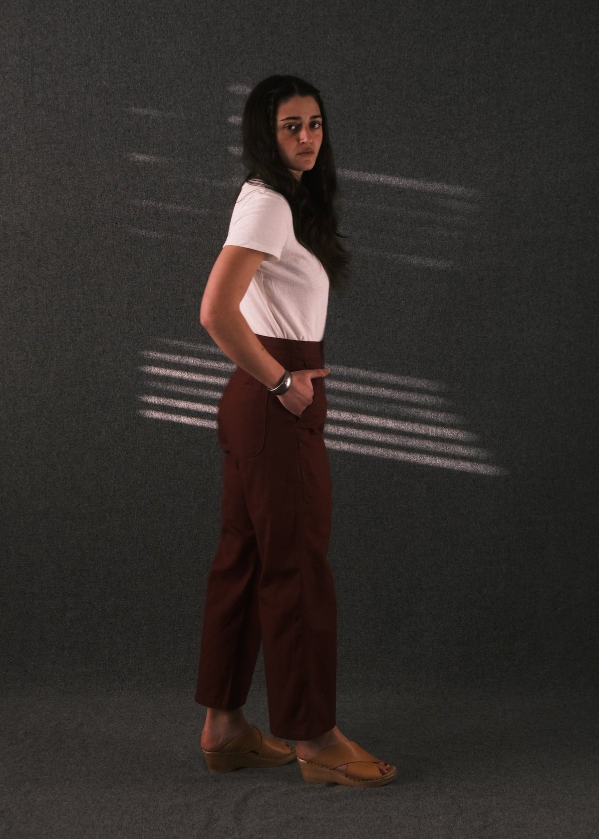 A woman stands in profile wearing a white short-sleeve top and dark brown wide-leg trousers against a dark gray studio backdrop with horizontal light streaks.