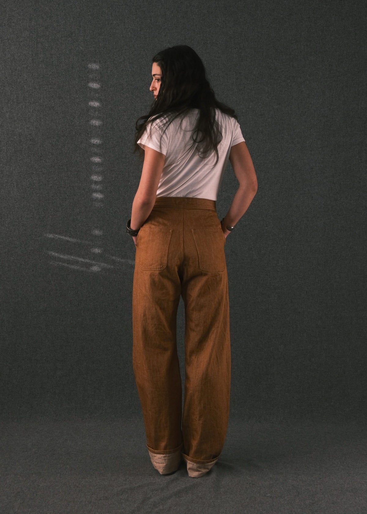 A woman is shown from behind wearing a white t-shirt and high-waisted, wide-leg brown corduroy trousers against a dark gray textured studio backdrop.