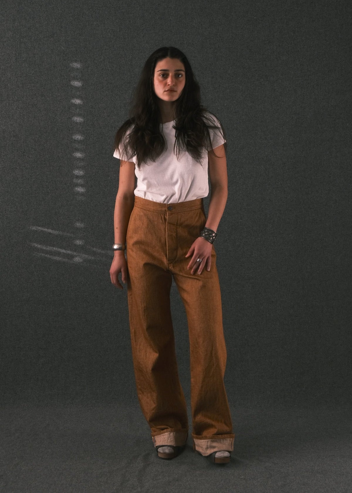 A woman stands against a dark gray textured studio background wearing a white short-sleeve t-shirt and high-waisted, wide-leg brown pants.