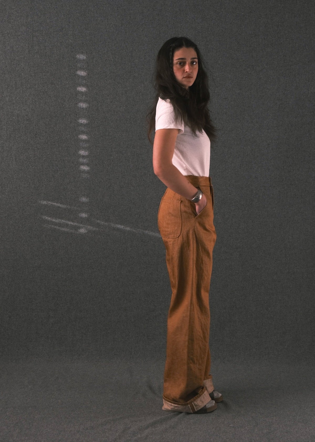 A woman stands in profile wearing a white short-sleeve t-shirt and brown wide-leg pants against a dark gray studio backdrop.