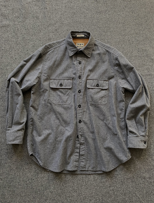 A gray long-sleeve button-up shirt with two chest flap pockets, laid flat on a textured gray background.