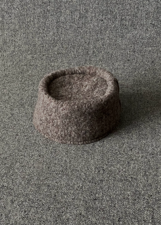 A small felt fez hat in a speckled taupe-brown color, photographed from a high angle on a textured gray carpet background.
