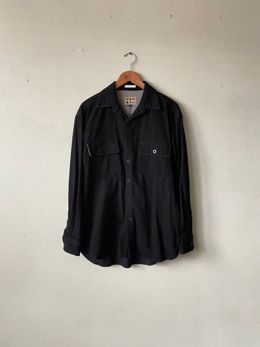 A black long-sleeve button-up shirt with two chest pockets, hanging on a wooden hanger against a plain, light beige wall.