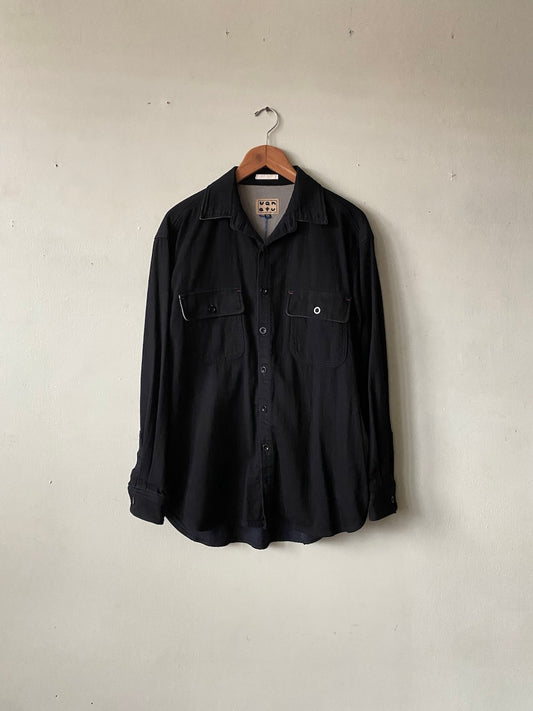 A black long-sleeve button-up shirt with two chest pockets, hanging on a wooden hanger against a plain, light beige wall.