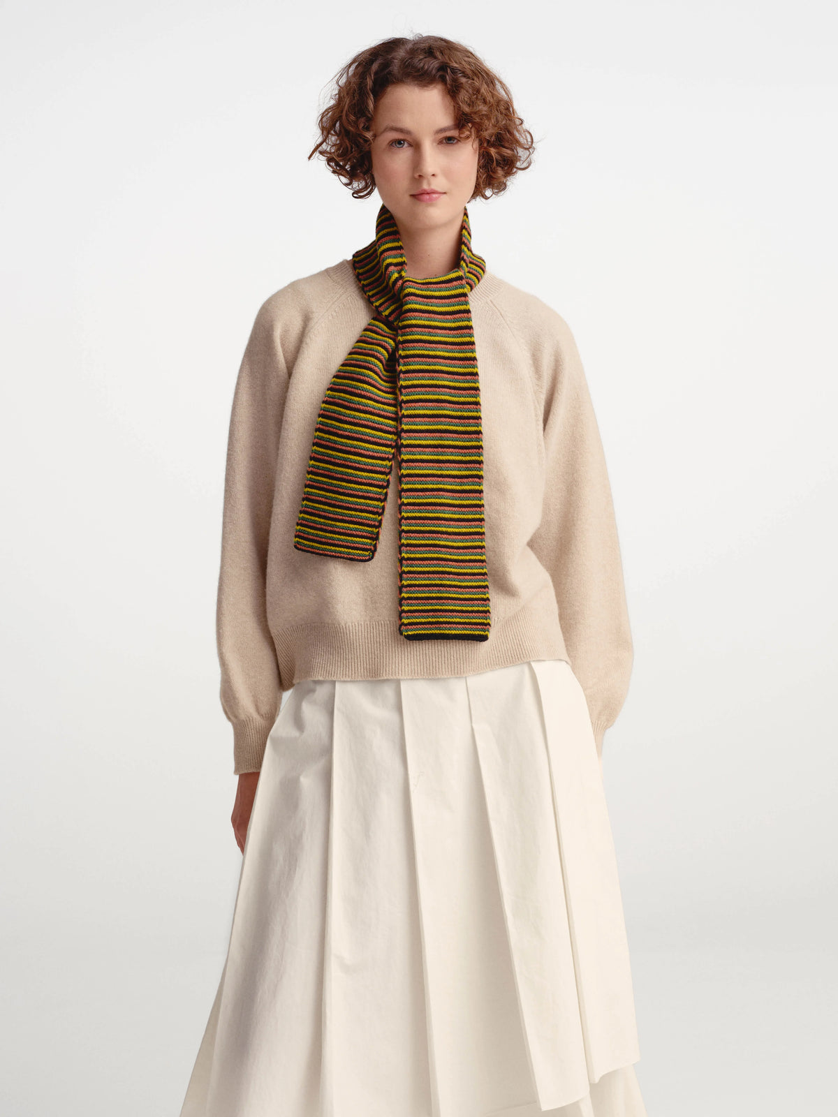 A model wearing a beige knit sweater, a pleated white skirt, and a striped knit scarf in olive green, yellow, and black, set against a plain light gray studio background.