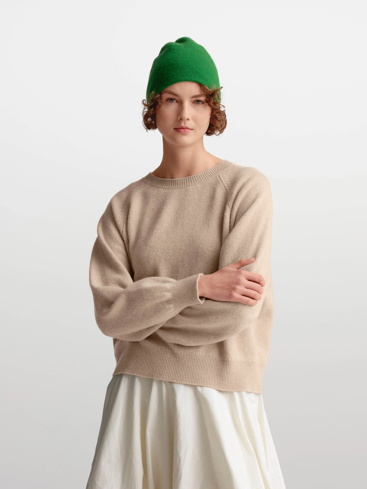 A model wearing a beige knit crewneck sweater, a green beanie, and an off-white skirt, standing against a light gray studio background.
