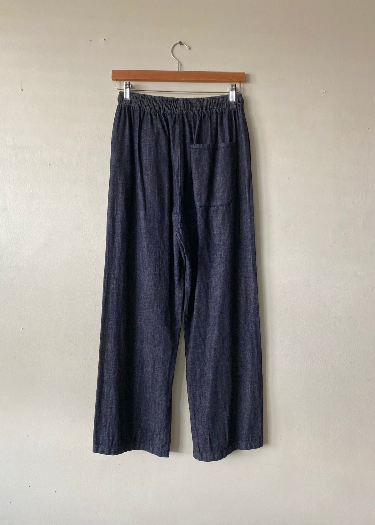 Dark indigo wide-leg cropped pants with an elastic waistband and a side pocket, hanging on a wooden hanger against a plain beige wall.