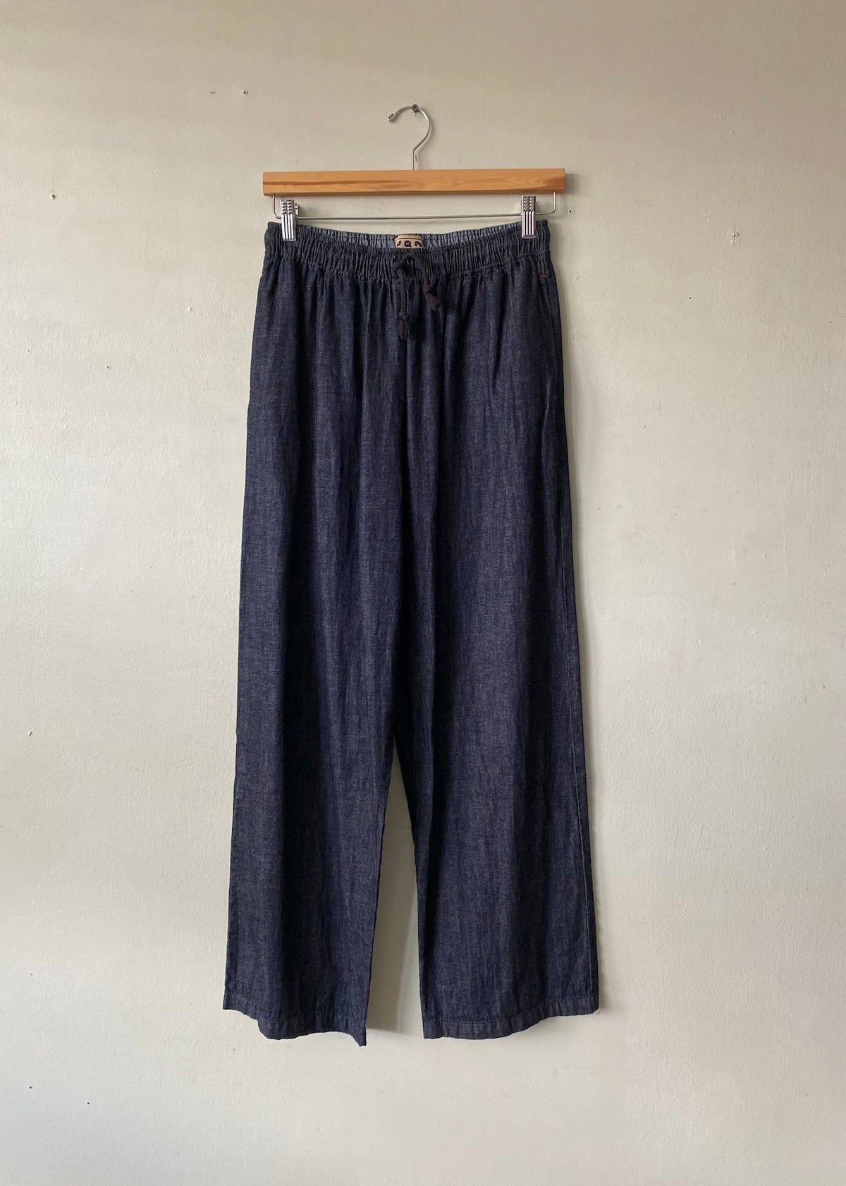 Dark indigo wide-leg pants hanging from a wooden hanger against a plain beige wall.