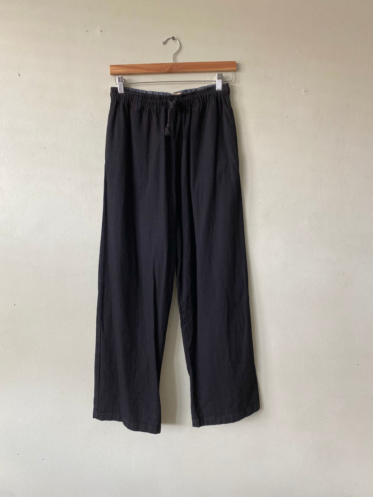 Black wide-leg cropped pants hanging from a wooden hanger against a plain off-white wall.