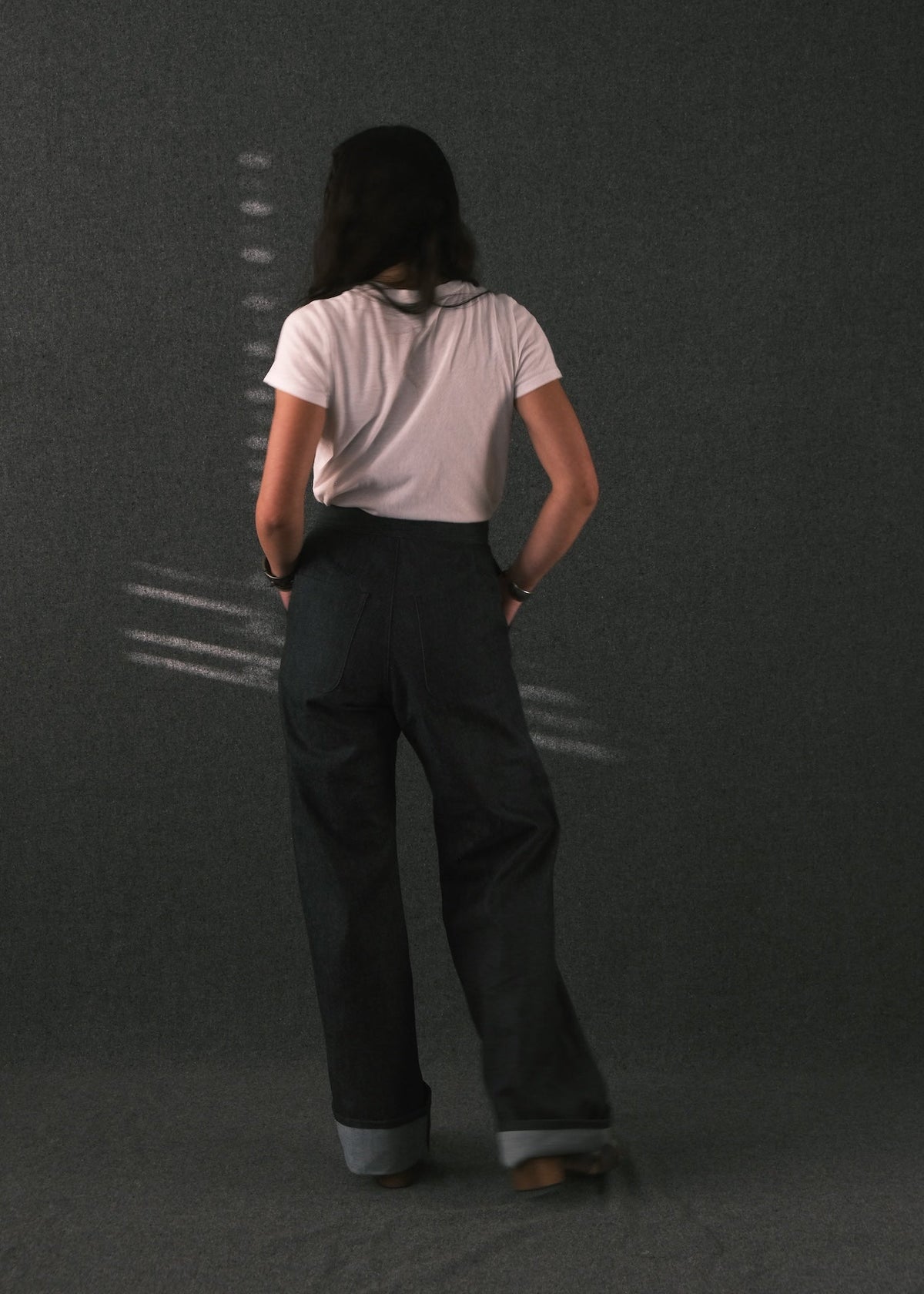 A person viewed from behind wearing a light pink short-sleeve t-shirt and dark charcoal wide-leg trousers with rolled cuffs, standing against a dark textured studio background.