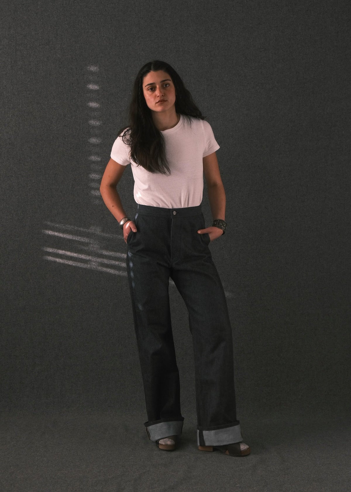 A woman stands against a dark gray textured studio backdrop wearing a light pink short-sleeve t-shirt and dark charcoal gray wide-leg jeans with cuffed hems.