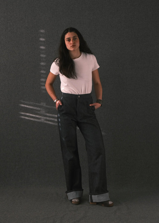 A woman stands against a dark gray textured studio backdrop wearing a light pink short-sleeve t-shirt and dark charcoal gray wide-leg jeans with cuffed hems.