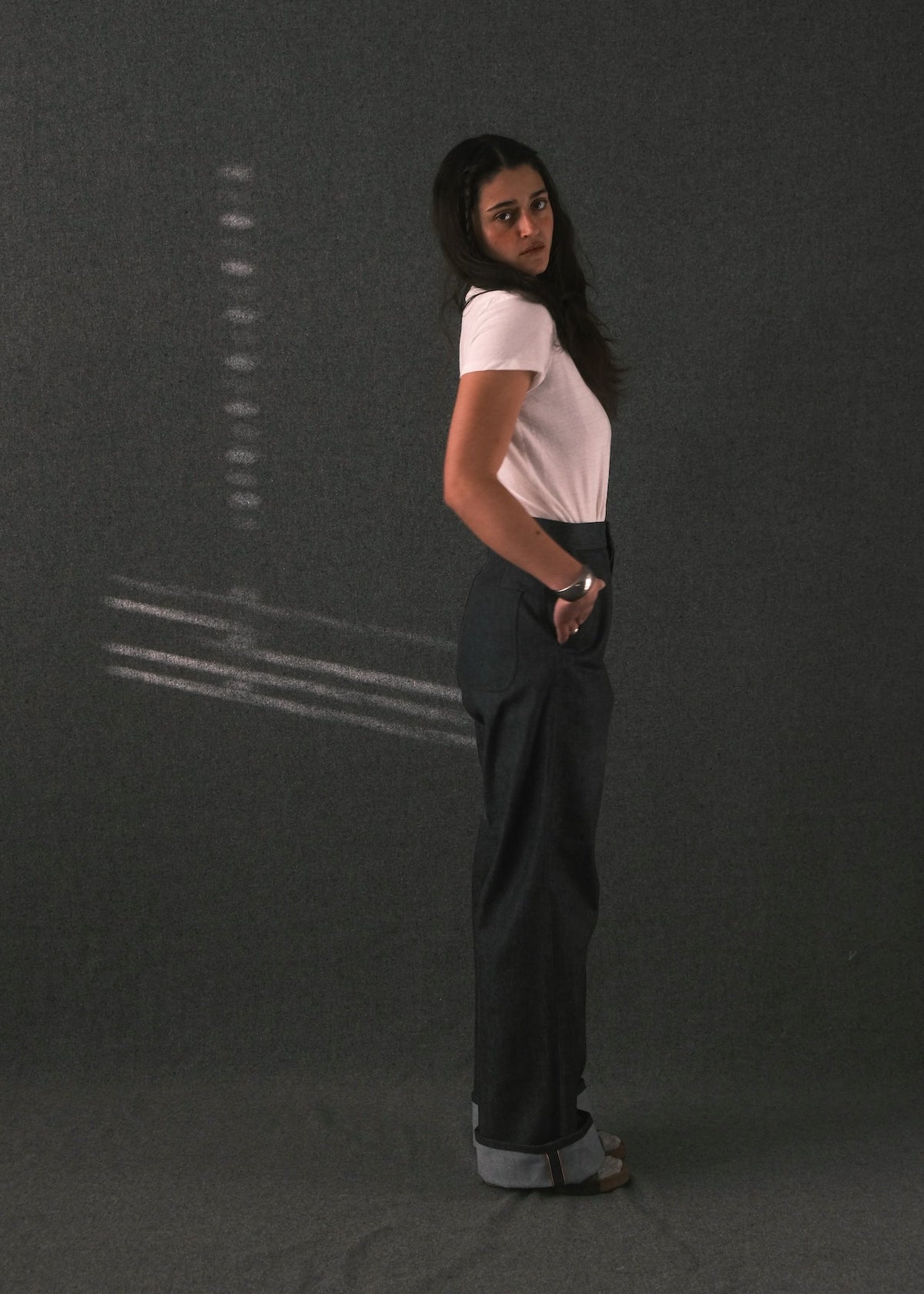 A woman stands in profile against a dark gray studio backdrop, wearing a light pink short-sleeve top and dark charcoal wide-leg pants.