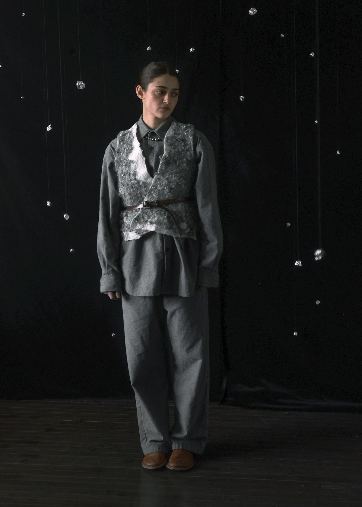 A person stands against a dark studio backdrop wearing a gray outfit with a patterned vest, dark shirt, and brown shoes.