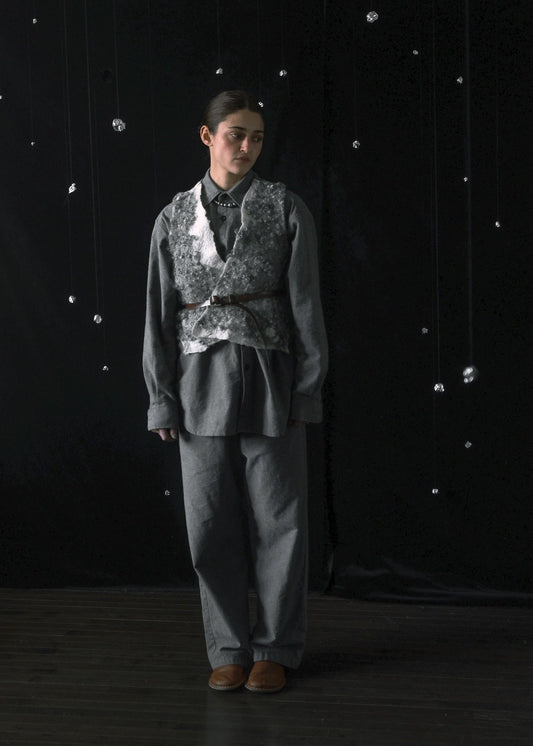 A person stands against a dark studio backdrop wearing a gray outfit with a patterned vest, dark shirt, and brown shoes.