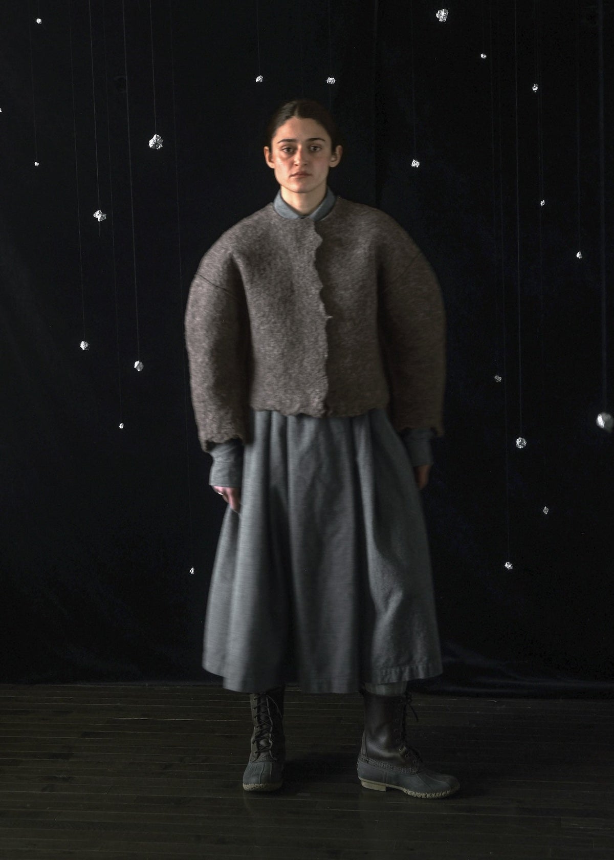 A model stands against a dark backdrop with hanging silver stars, wearing a textured taupe cropped cardigan over a gray dress and dark boots.