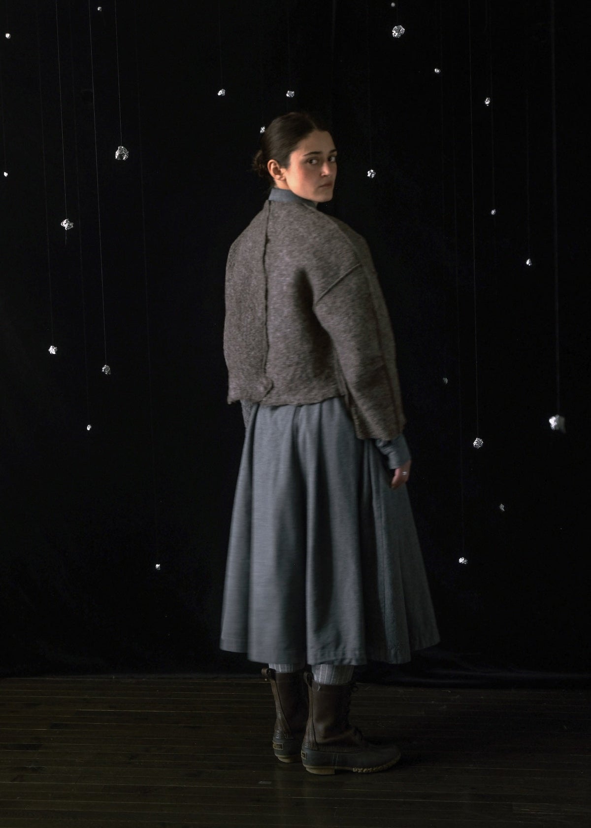 A model stands in profile wearing a textured gray cropped jacket over a blue-gray skirt, set against a dark studio backdrop with hanging reflective spheres.
