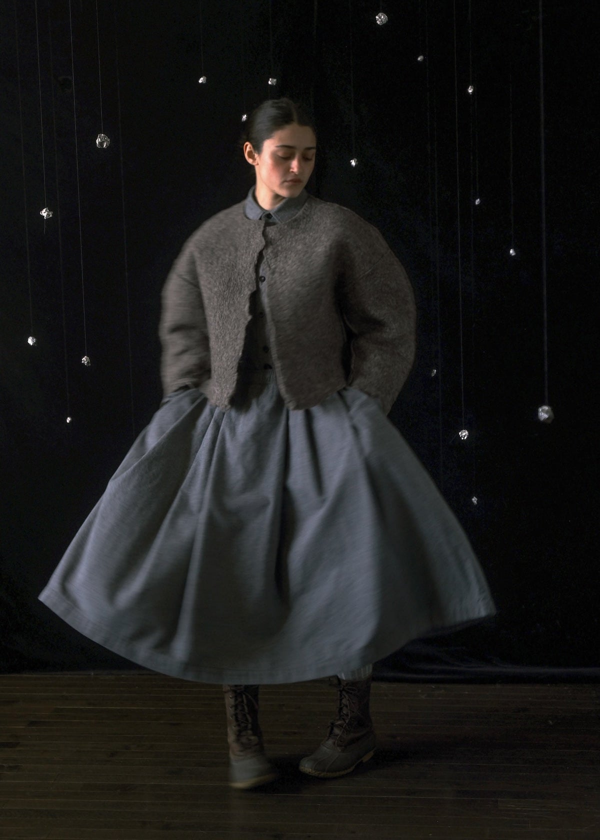 A model stands in a studio wearing a voluminous gray skirt dress with a textured taupe-gray cropped cardigan and dark lace-up boots. The background is a dark black curtain decorated with hanging silver glass beads.